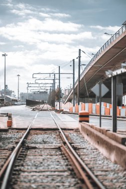 Şehir demiryolu bir sanayi limanına doğru gidiyor. Tepede elektrik kabloları, beton bariyerler ve bir otoyol köprüsü bulunuyor. Sahnede metal direkler, yol işaretleri ve bulutlu bir gökyüzü var.