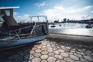 Kırık bir taş iskeledeki kırık camları ve paslı metal parçaları olan terk edilmiş ve çürümüş mavi bir balıkçı teknesinin geniş açılı görüntüsü. Arka planda vinçleri, tekneleri ve şehir silueti olan sanayi limanı