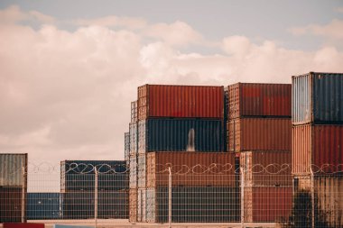 Endüstriyel bir deponun dikenli tellerinin arkasında kırmızı ve mavi renkli konteynırlar yığılmış. Konteynırlar bulutlu bir gökyüzünün altında pas ve aşınma izleri gösteriyor.