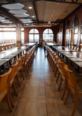 Uzun ahşap masalar ve bir grup etkinliği veya ziyafete hazırlanan sıralar, kemerli pencerelerden yansıyan sıcak güneş ışığı, geniş açılı dikey çekim