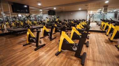 Modern kapalı bisiklet stüdyosu ahşap zeminde birden fazla sarı sabit egzersiz bisikletleri aynalı duvarlar, tavan ışıkları ve eğitmen rehberliği için bir platform.