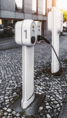 Çağdaş elektrikli araç şarj istasyonu ofis binası yakınındaki kaldırımda, parlak beyaz tasarım ve doğal gün ışığında fişe takılmış siyah kablo; dikey geniş açılı çekim