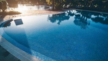 Sakin mavi su, fayans detayları, tropikal palmiye yansımaları ve arka planda güneşli şezlonglar ile lüks bir tatil köyünde sabah erken sahnesi; Punta Umbria, İspanya