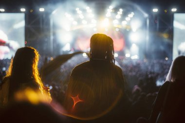 Parlak sahne ışıkları, duman ve merceklerin oluşturduğu canlı bir gece konserinin keyfini çıkaran insanların siluetleri rüya gibi bir atmosfer yaratıyor. Önplan figürlerine seçici odak, alanın sığ derinliği