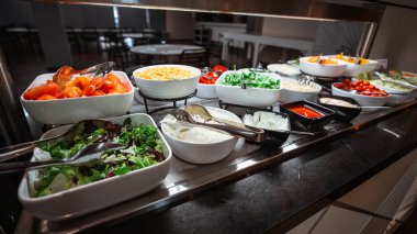 Taze ve renkli salata barı, domates, marul, mısır, soğan, roka, sos ve malzemeler, beyaz seramik kaseler gibi çeşitli sebzelerle servis edilen bir açık büfe.