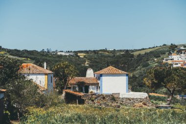 Geleneksel beyazlatılmış evler, terasotta çatıları, mavi süslemeler, taş duvarlar ve parlak mavi gökyüzünün altındaki yemyeşil tepeler ile Aldeia da Mata Pequena 'nın resimli kırsal manzarası.