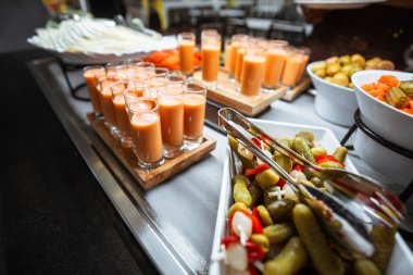 Turşulu sebzeler, taze havuç ve salatalık dilimleri ile renkli büfe masası ve kremalı gazpacho ya da sebze püresi ile dolu sıra sıra küçük bardaklar, self-servis için hazır.