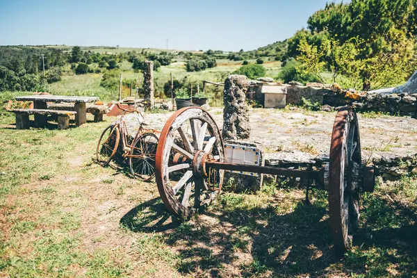Parlak bir yaz gökyüzü altında taştan banklar ve yeşil yuvarlanan tepeler ile çevrili, eski ahşap bir araba aksı ve yıpranmış bisiklet ile kırsal manzara; Aldeia da Mata Pequena