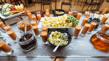 Renkli kahvaltı ya da açık büfe masasında çeşitli peynirler, taze meyveler, kurutulmuş meyveler shot bardakları, reçeller ve bal içinde mini içecekler, hepsi güzel bir şekilde düzenlenmiş parlak ve zarif bir ortam.