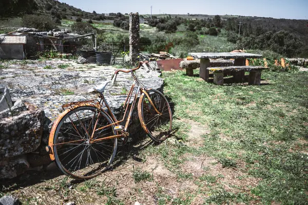 Terk edilmiş paslı bisiklet, kırsal bir alanda yıpranmış taştan bir duvara yaslanmış, yosun kaplı piknik masası ve parlak bir yay gökyüzünün altında yeşil tepeler.