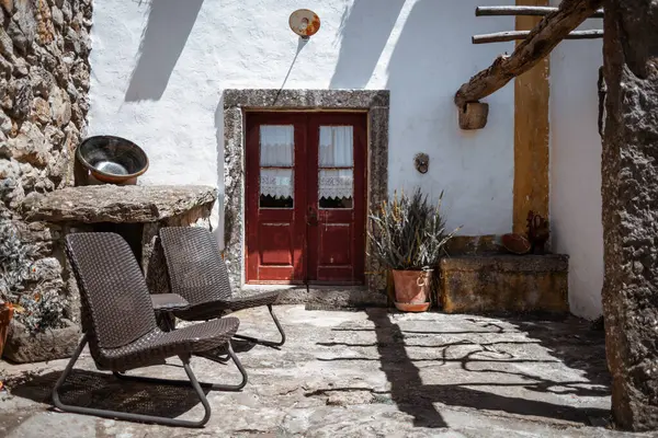 Aldeia da Mata Pequena 'da iki hasır sandalye, yıpranmış bir taş masa, dantel perdeli kırmızı ahşap kapılar, saksı bitkileri ve ılık doğal ışık gölgelerinin yer aldığı rahat bir köy avlusu.