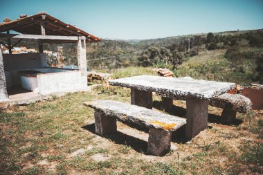 Yıpranmış taş piknik masası ve kırsal bir alanda banklar, yakınlarda tepeler ve açık mavi gökyüzünün altında bitki örtüsüyle çevrili geleneksel bir açık hava yıkama yapısı.