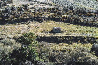 Telefoto merceğiyle çekilen kırmızı kiremit çatılı izole edilmiş küçük bir taş kulübe. Uzak mesafede yoğun çalılıklar ve tarlalarla çevrili, güneşli bir teras arazisine yerleştirilmiş.
