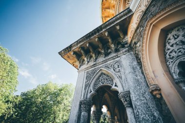 Süslü taş kemerler, karmaşık oymalar ve Berberi etkileriyle, parlak mavi bir gökyüzüne ve yemyeşil ağaçlara karşı kurulmuş, büyük tarihi bir yapının alçak açılı mimari detayları.