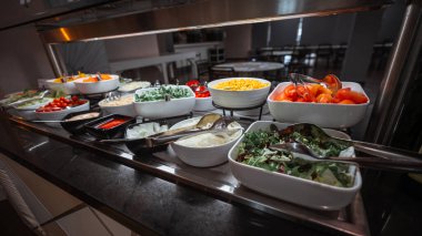 Domates, mısır, soğan, marul, roka, vişneli domates ve soslar dahil olmak üzere çeşitli taze sebzelerle dolu renkli salata büfesi, sıcak ışık altında beyaz kaselerde sergileniyor.