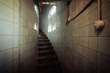 Dar fayanslı merdiven boşluğunun geniş açılı iç manzarası kemerli pencerelerden yukarı doğru doğal ışığa doğru kıvrılıyor. Açığa çıkmış borular ve karamsar gölgeler atmosferik, sinematik bir his yaratır.