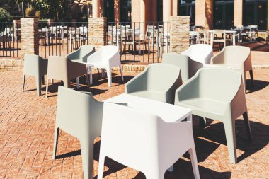 Güneşli bir terasta beyaz masaların etrafına dizilmiş modern plastik koltukları olan açık hava oturma alanı tuğla döşeme ve arka planında zarif bir taş ve metal çit var.