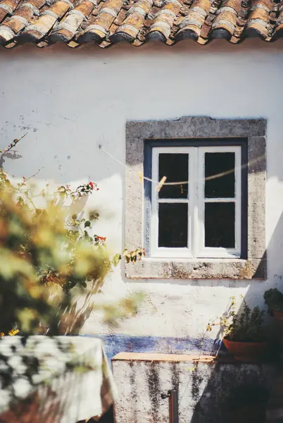 Taşla çerçevelenmiş küçük ahşap pencereli, kırsal beyazlı duvar, güneş ışığıyla kaplı terasotta kiremit çatı altında, tırmanma tesisleri ve rahat bir açık hava oturma köşesi ile çevrili.