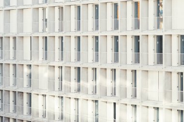 Minimalist beyaz balkonları, cam kapıları ve tekrarlayan geometrik mimarisi olan, yumuşak doğal gün ışığıyla aydınlatılan ve ince gölgeler bırakan modern bir apartman veya otel binasının yan görüntüsü.