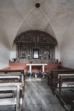 Camino de Santiago boyunca küçük bir köy şapeli (capilla), içinde dini figürler, ahşap banklar ve taş zeminde çiçek sunan oyulmuş ahşap bir sunak bulunur.