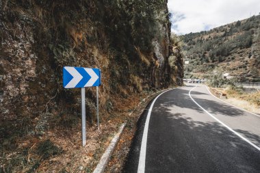 Keskin virajlı dağ yolu, kayalık arazi ve orman yamaçlarıyla çevrili mavi bir sembolle çevrili, parçalı bulutlu bir gökyüzü altında seyahat veya yol güvenliği temaları için mükemmel.
