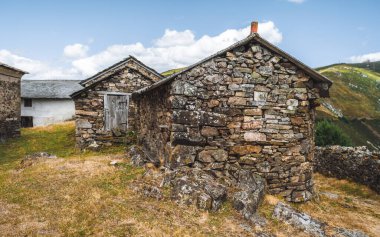 Dağlık bir kırsalda eski taştan kulübelerle kaplanmış ahşap kapılar, kuru çimenler, kayalık arazi ve yuvarlanan yeşil tepeler yumuşak mavi bulutlar altında.
