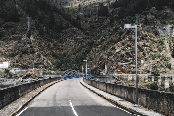 Beton bir baraj boyunca uzanan yol, mavi baraj kapaklarıyla çevrili, sarp kayalık yamaçlar, elektrik hatları ve seyrek bitki örtüsü, bulutlu bir dağ manzarası altında.