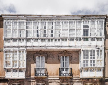 Geleneksel Galiçya binası cephesi. Süslü siyah demir balkonları, yıpranmış taş duvarları ve parlak bulutlu gökyüzünün altında geniş beyaz ahşap galeri pencereleri var.