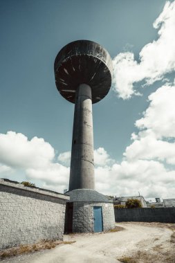 Parlak mavi gökyüzü altında, şehir altyapısını ve endüstriyel mimariyi sembolize eden, dağılmış bulutların altında yükselen yüksek beton su kulesi.