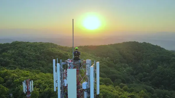 Technician working on the top of a Telecom tower, sunset in the mountains. Aerial