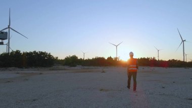 Günbatımında rüzgar türbini çiftliğinde yürüyen teknisyen mühendisi yenilenebilir temiz yeşil enerjinin verimini kontrol ediyor