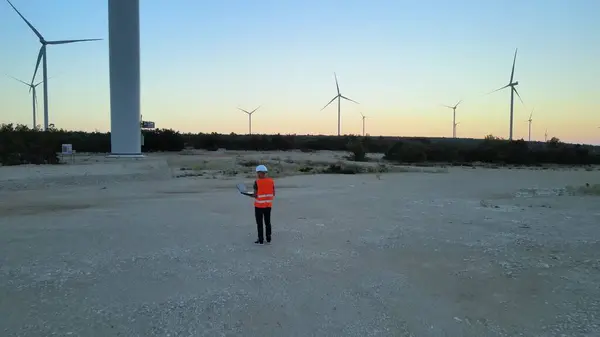 Aerial of engineer with laptop inspecting and control the efficiency of windmill wind turbine farm at sunset, clear renewable green energy zero emission concept