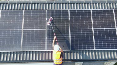 Çatıdaki fotovoltaik güneş panellerini paspas kullanarak temizleyen üniformalı ve miğferli bakım teknisyeni. Sürdürülebilir yenilenebilir yeşil, biyolojik ve ekolojik temiz enerji