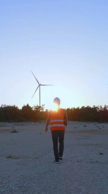 Günbatımında yel değirmenine doğru yürüyen mühendis. Rüzgar Türbinleri Teknoloji Elektrik Mühendisliği Konsepti. Dikey yönelim