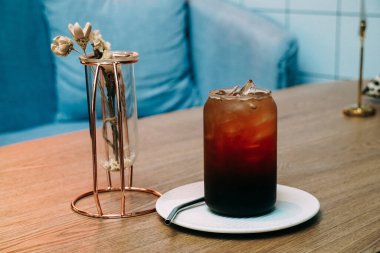 Tahta masanın üzerinde saman olan bir fincan soğuk buzlu kahve. Bir bardak buzlu americano kahvesi, ahşap masa ve mavi kanepe arka planı, seçici odak.