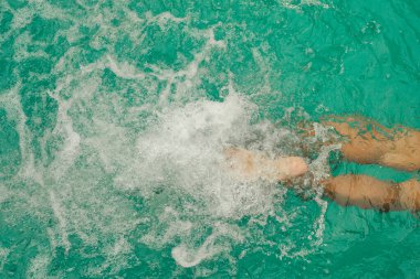 Adamın bacakları yüzme havuzunda. Mavi suda yüzen adamın alt kısmının üst görüntüsü. Ayaklar yüzme havuzunda su sıçratıyor. Tatil ve yaz tatili