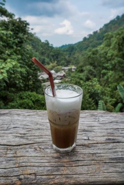 Açık havada buzlu kahve bardağının yakın çekimi. Kafe Latte, gözlüklü, uzun boylu, tahta tabakta, doğal yeşil manzaralı. Mae Kampong köyündeki kafede buzlu kahve, Chiang Mai, Tayland.