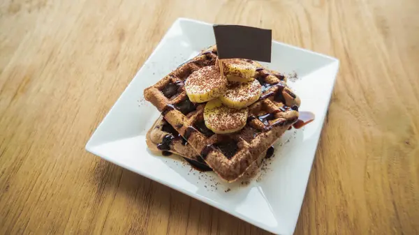 Breakfast waffles with bananas and chocolate sauce. Traditional waffles with fresh banana and Chocolate sauce. Wood background. Top view