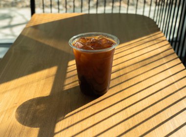 Buzlu kahve. Ahşap masa arkasında cam bardakta. Pencereden gelen güneş ışınıyla. Yaz ferahlatıcı içecek. Sabah sade kahve, güneş ışığı, kafede bir bardak americano..