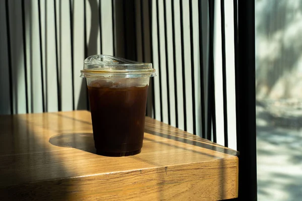 Coffee with ice cubes in drinking glass on wood table background with a sunbeam from the window. Summer refreshing drink. Black coffee in the morning with sunlight, a glass of americano in the cafe.