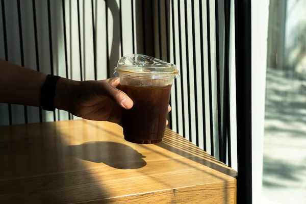 Hand holding black ice Coffee in drinking glass on wood table background with a sunbeam from the window. Summer refreshing drink. Black coffee in the morning with sunlight.