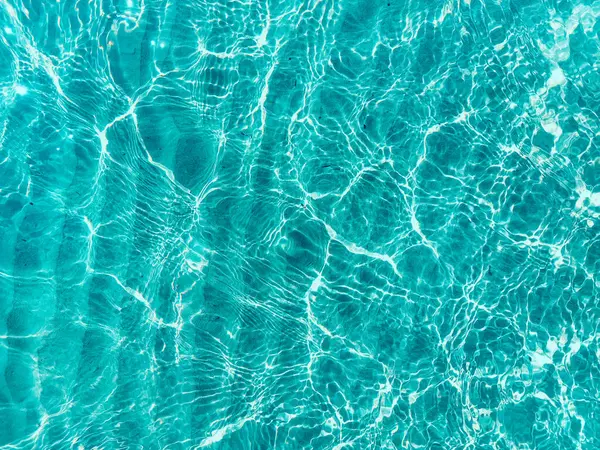 Water ripple over sandy beach. Transparent blue colored clear water surface texture with splashes. Water background, ripple and flow with waves. Blue water shinning. Sea, ocean surface. Overhead top view. Flat lay design.