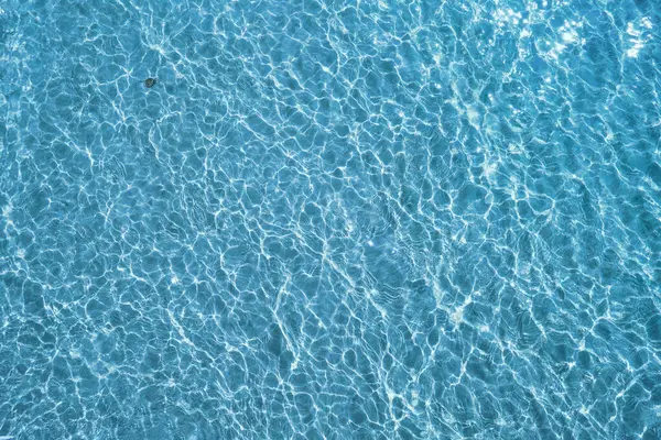 Water ripple over sandy beach. Transparent blue colored clear water surface texture with splashes. Water background, ripple and flow with waves. Blue water shinning. Sea, ocean surface. Overhead top view. Flat lay design.