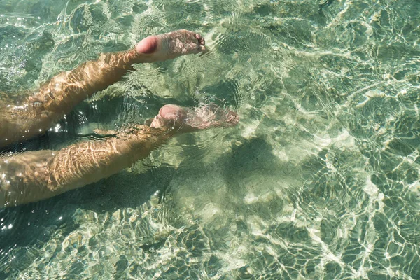 Female legs in crystal clear water on a beach with sandy. Legs dipped in clear water with blue bottom on sunny summer day. Woman feet swim underwater beneath the surface swimming under the crystal clear blue water in the sea.