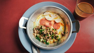 Kahvaltı menüsünde sosis, Çin sosisi, jambon, kıymalı domuz eti ve doğranmış soğandan oluşan sıcak tavayla servis edilen tavada kızartılmış yumurta var..