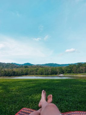 Piknik paspasında çıplak ayaklı kadın bacakları yeşil çimenler göle ve dağlara bakıyor. Kadın güneşli bir günde Jedkod Park, Tayland 'da dinleniyor.