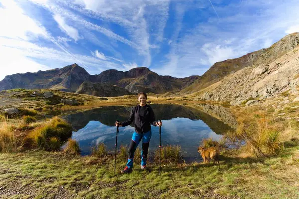 latina woman with her dog in a mountain lake where clouds and mountains are reflected, with a group of mountain hikers behind her