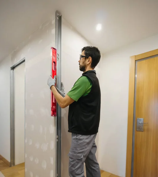 professional handyman works on a plasterboard wall in an apartment with his level, glasses and work clothes.