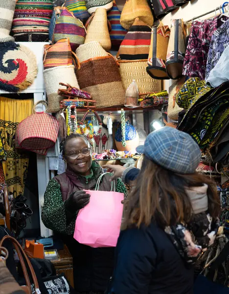 older black african woman from nigeria selling handbags and handicrafts in her store to a customer. pink bag