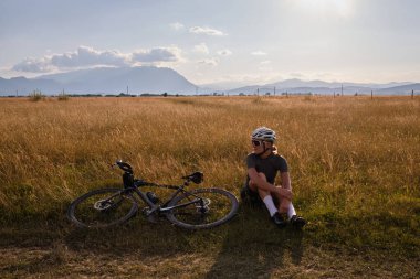 Bisikletli, yeşil formalı, siyah şortlu ve kasklı bir kadın dağdaki bisiklet antrenmanı sırasında dinleniyor. Yol bisikleti kavramı. Gün batımında bisiklet süren kadın..